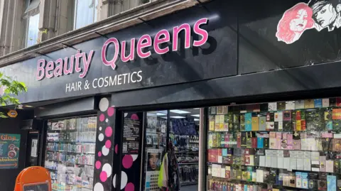 The front of a shop has a sign saying "Beauty Queens" and there are lots of items lined up inside the front windows on either side of the entrance door.