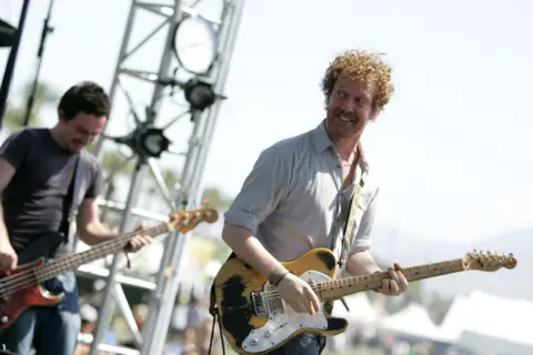 Getty Images Two men on stage playing with a band, the man on the left is holding a yellow and white electric guitar and turning his head back towards a bandmate. He has an amazed smile on his face. Behind him is a man playing a bass guitar, he too is smiling while looking down at the instrument he is playing. It is daylight at a music festival. 