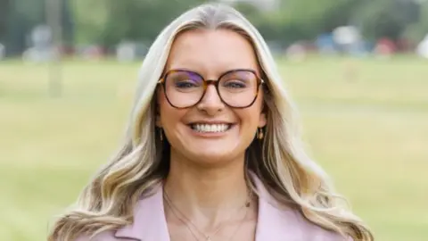 A woman with long blonde hair and glasses smiles while looking directly at the camera. She is wearing a pink jacket, with a green park in the background 