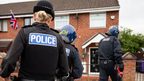 Three police officers taken from behind in riot gear one holding a battering ram as they arrive at a house to execute a warrant