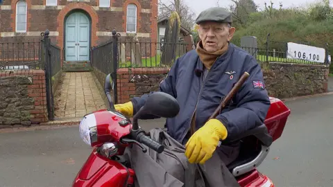 91-year-old Terry Meakin is sitting on his red mobility scooter.  He is wearing a grey flat cap, a brown scarf, a blue bomber jacket and yellow gloves. 