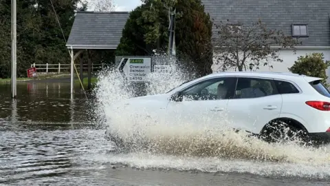 BBC A white car makes its way through the flood waters in Purton, Wiltshire