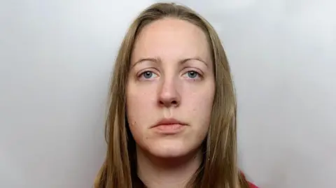 Lucy Letby police mugshot. Sandy blonde hair is worn long over a red hoody. She has thin eyebrows and bluey grey eyes.