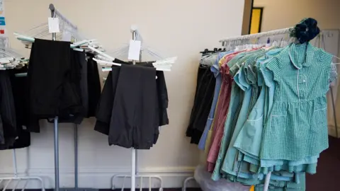 Clothing racks of school uniform, with black trousers hanging on the left, and a rail of green gingham summer dresses on the right.