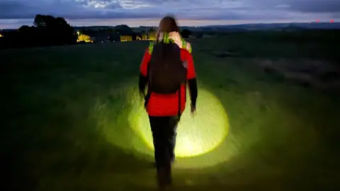 A walker strides in a field in Northumberland. The light from their head torch illuminates the grass beneath their feet. They are dressed in an orange t-shirt which has the logo of the Motor Neuron Disease Association.