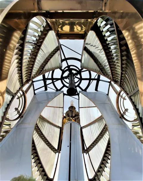 Victor Tregubov A view from inside a lighthouse lantern room, showing the curved glass Fresnel lenses and metal framework surrounding the central light.