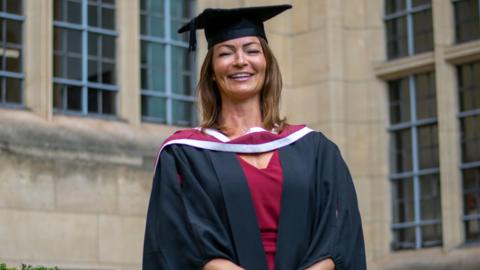 Dr Becs Bradford pictured at her university graduation. She is wearing a red dress with a black graduation gown over the top. She is also wearing a graduation cap. She is pictured smiling at the camera