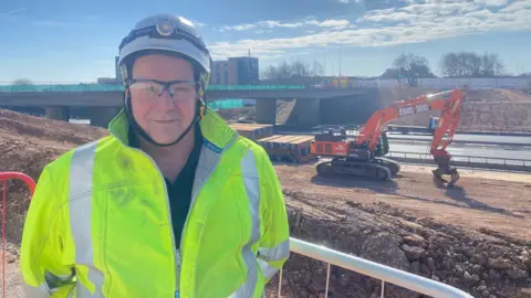 BBC Terry Robinson in a high vis jacket and white helmet. He is smiling at the camera, and there is a digger and the bridge in the background. 