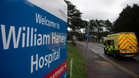 A hospital sign next to a road with an ambulance driving past it. The sign reads "William Harvey Hospital".