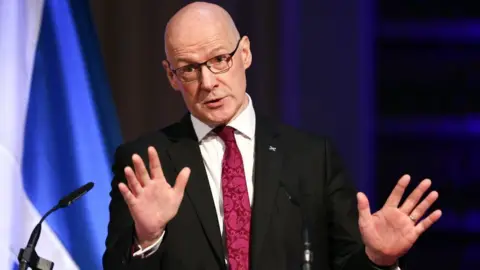 PA Media A bald man with glasses, wearing a dark suit and pink tie, stands with his hands raised in front of his body as he speaks at a podium. A saltire is illuminated in the background.