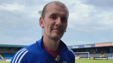 A man looks into the camera. He is wearing a blue tracksuit top with the Glenavon badge and white stripes on the sleeves, and a red t shirt. He has balding brown hair. A small football stand with blue seats and a football pitch is in the background.
