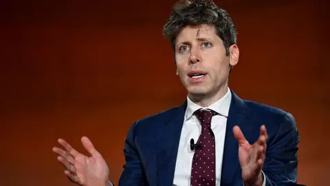 Bloomberg via Getty Images An image of Sam Altman speaking. He has blue eyes and short dark brown hair, and is wearing a navy blue blazer with a white shirt underneath and a burgundy tie with small white polka dots. He is gesturing with both his hands addressing a crowd off camera, behind him is a blurred copper background 