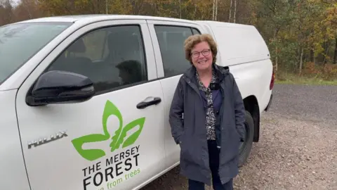 BBC Professor Eunice Simmons has short brown hair and is wearing dark rimmed glasses and a blue coat and is stood next to a white van with The Mersey Forest logo on.