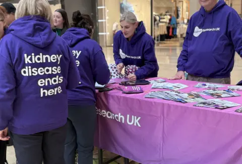 A stall in Queensgate Shopping Centre which is covered in pink fabric and has piles of leaflets fanned out on its top. A woman in a purple hoody saying Kidney Research UK is leaning across the table to another couple of women, facing her in purple hoodies which say on their backs kidney disease ends here. 
