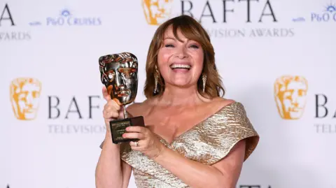 Lorraine Kelly smiling in a gold dress while holding a Bafta television award