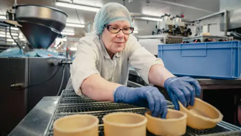 William Sword A woman in a bakery production centre, leaning over to adjust pastry 