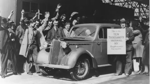 Vauxhall Heritage A black and white picture of an old car coming off the production line at Vauxhall Motors in Luton. There are people around it with their arms in the air and a man wearing a suit and hat who is holding a placard which reads: "This is the ten thousandth Vauxhall 10 bought in under five months".