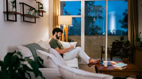 Getty Images A man with brown hair and beard wearing a green top and white pants writing on a notepad sat on his sofa. 