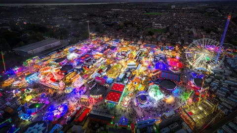 PA Media Una vista aérea de una feria de atracciones adornada con luces de colores de neón que rodean una ciudad por la noche. Puedes ver una noria, atracciones altas que parecen postes, remolinos y estrellas.