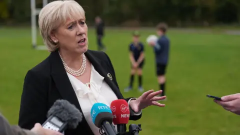 PA Heather Humphreys has short blonde hair and is dressed in a black jacket and white blouse. She is gesturing with her left had and standing beside broadcast media microphones. In the background people can be seen on a football pitch.