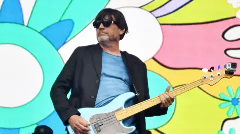 Jim Dyson/Getty Images A man wearing a black blazer and sunglasses while playing a blue bass guitar.