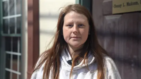 Joy Coker looks directly at the camera, wearing a white hooded sweatshirt with her hair down and blowing in the wind