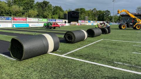 Parts of the artificial pitch rolled up and being lifted away by machines