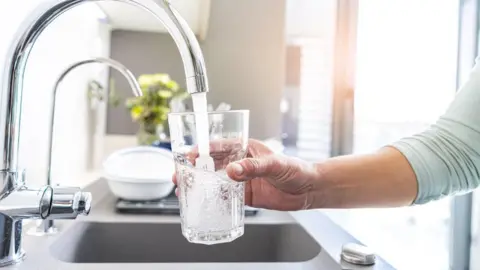 A person's hand is seen holding a glass under a running tap and filling it with water in a kitchen