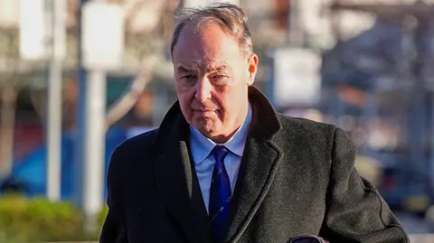 PA Media Carley, who has silver hair, walking wearing a suit, blue shirt and blue tie and a long dark coat past some railings with a bag under his left arm