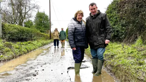 A man and woman, both wearing Wellington boots and dark coats, standing in a deep pothole on a country lane, with green bushes either side. Two people can be see standing on the road in the background.