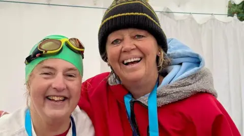 Liz Davey Two women in their 60s, Liz Davey and Jacqui Hanlon, stand close together and are laughing. One has a green hat and googles and is wearing a white top, the other has a black and yellow bobble hat and is wearing a dry robe. They both have medals round their neck. 