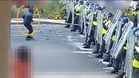 Northumbria Police Kennedy in black pushes a large metal pole towards a line of police officers in riot gear with shields. 