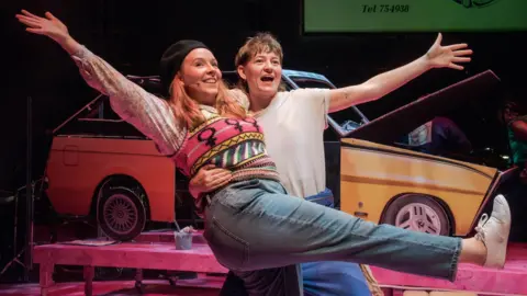 Chris Saunders Two performers on stage in a playful pose, with one lifting the other who has arms outstretched and one leg raised. Behind them is a yellow car with its hood open, along with stage props including a bench and an old-fashioned telephone, suggesting a theatrical performance with vibrant lighting.