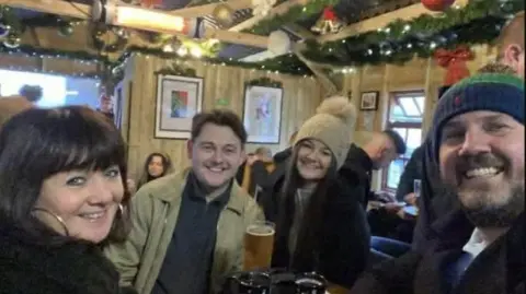 Charlie Hurst a group of four people in a pub, a pint can be seen and all four are smiling - wrapped up in winter clothes. Christmas decorations can be seen in the background.