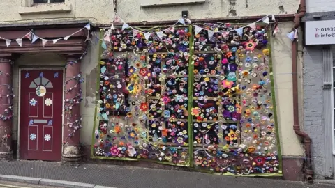 A derelict cream coloured hotel with a sign that reads "white horse hotel". On its windows there are hanging cascades of dozens of colourful knitted flowers. there are knitted vines and flowers entwined around pillars, and flowers stuck on the door.