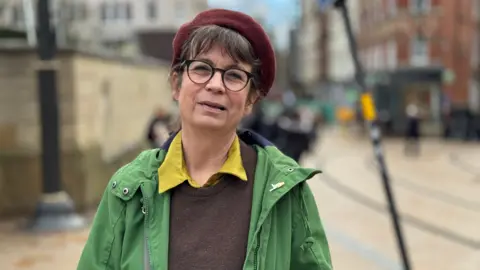 A woman wearing a green coat and red hat stands in the middle of Birmingham city centre.