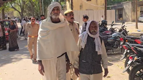 Two men wearing white scarfs and traditional Indian outfits walking with policemen who are wearing uniform