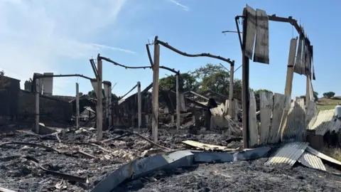 Pictured is a huge pile of twisted steel beams and collapsed corrugated steel panels. Four steel frames are just about still standing, giving you a sense of the size of the barn. The metal is blackened and covered in dust. The sky is bright blue and it's clearly sunny. 