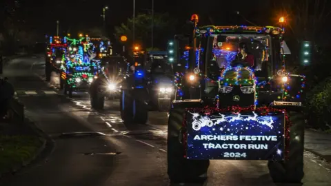 Five tractors one behind the other driving down the road at night. They all have colourful lights and Christmas decorations on them.