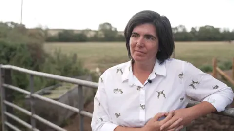 Abi Reader stands by a farm gate overlooking fields and countryside during an outdoor interview in rural Wales.