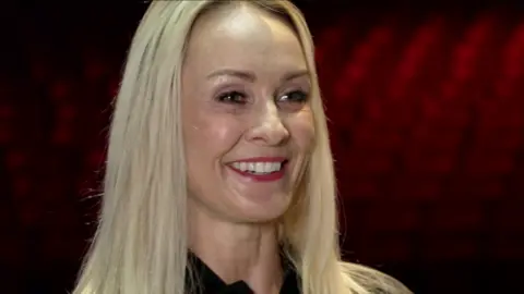 Laura Miller is smiling off camera to the right. She is wearing a black collared t-shirt. She has pink lipstick on and has blond hair.