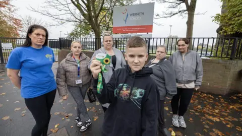 LDRS A picture of student Finlay holding up his sunflower lanyard in protest with some parents outside Werneth School on Harrytown road in Stockport