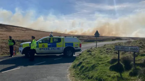 Side view of a West Yorkshire Police vehicle parked across a moorland road with two officers standing beside it. In the background a plume of brown and white smoke drifts across the moorland, partially obscuring an otherwise blue sky. 