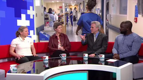 Four people sit on a red curved sofa on a TV set with a black curved table in front of them. Behind them is a large screen showing nurses and doctors working in a hospital.