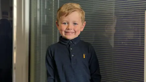BBC A young boy with dark blonde hair is smiling at the camera. He is wearing a dark navy shirt with a yellow logo on the breast in the shape of a stag's head