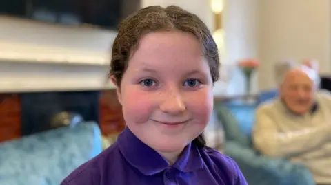 Helen Mulroy/BBC Eliza looks directly at the camera, she has brown hair pulled back in a pony tail and wears a purple school polo shirt