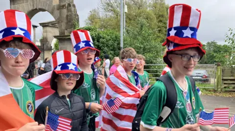 BBC Shows young footballers in green shirts, carrying Stars and Stripes flags and wearing Stars and Stripes hats