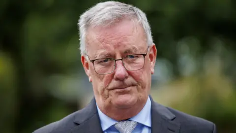 Mike Nesbitt looking at the camera, he has white hair and black glasses. He is wearing a dark suit and blue shirt with white and silver tie.