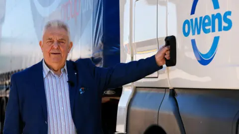 Huw Owen, wearing a blue blazer and striped shirt, stands next to the cab of one of his branded Owens Group lorries. He is holding the door handle and smiling at the camera on a sunny day.