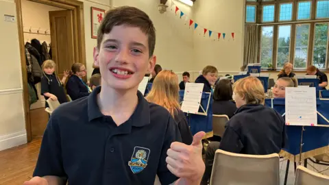 Joshua is wearing a navy blue shirt and giving thumbs-up. He is smiling. He is standing in a practice room with other brass players in the background.